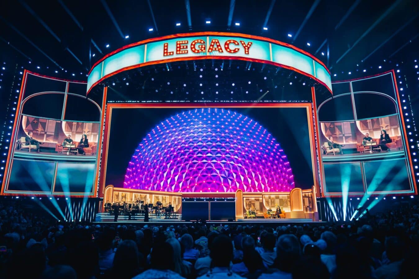 Broad stage setup with a curved LED sign reading LEGACY a glowing purple geodesic dome and audience in the foreground
