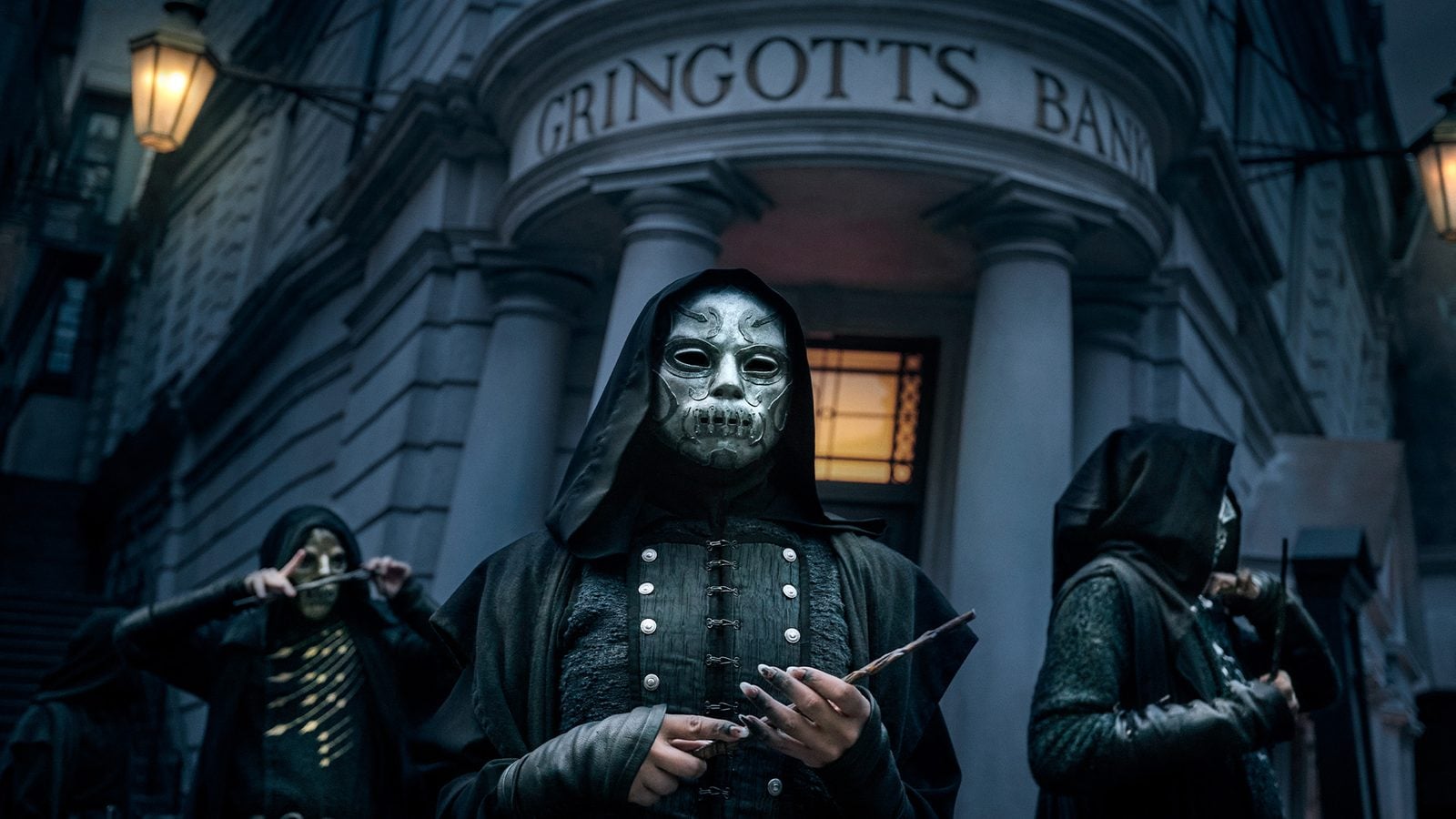 Hooded figure wearing a metallic skull mask stands before Gringotts Bank at night with other masked figures in dark cloaks nearby on a dim street