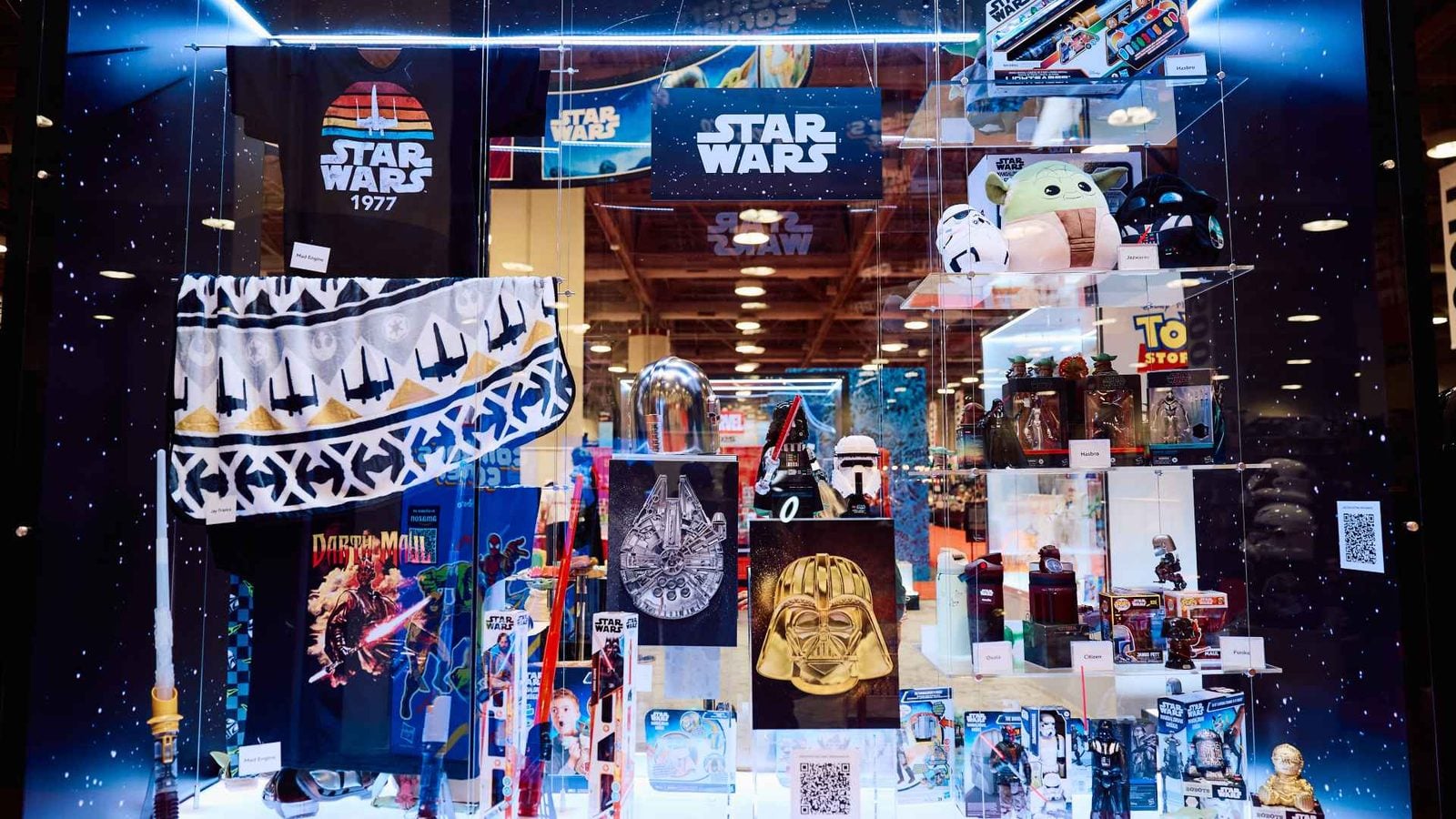 Star Wars store window display with plush Baby Yoda helmets figures posters and Tshirts arranged on glass shelves in a galaxy themed setup