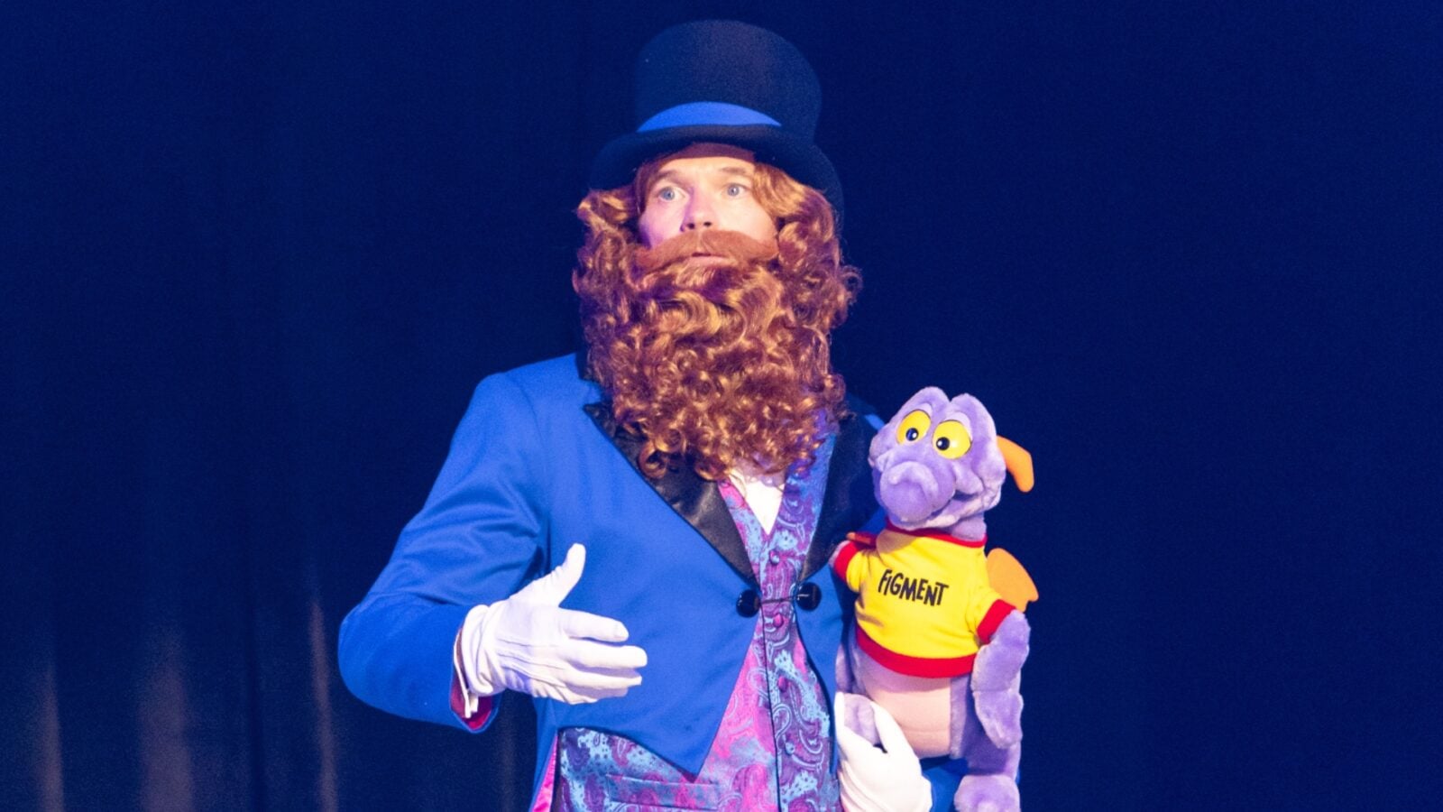 Stage performer in a blue suit and top hat with a curly red beard holding a purple dog puppet in a yellow FIGMENT shirt against a dark blue backdrop