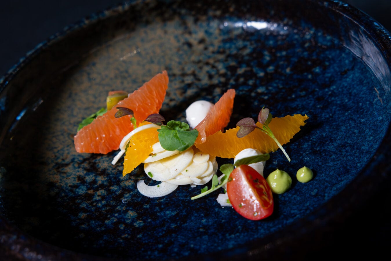 Close up of a plated dish with orange segments dollops of cream greens and a cherry tomato on a dark blue plate