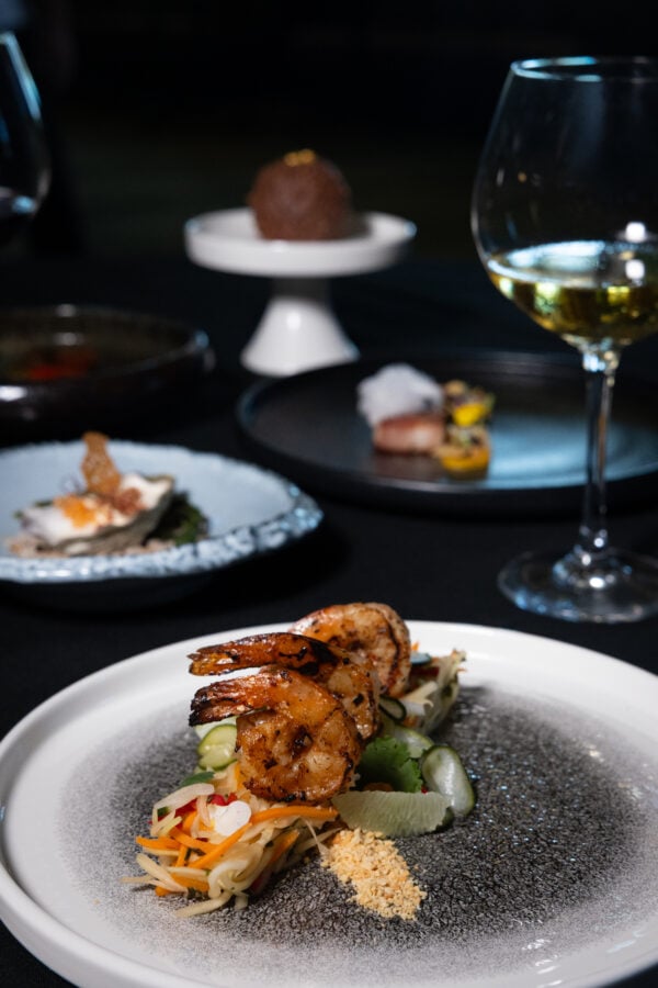 Plated shrimp appetizer atop a vegetable salad with sesame crumble on a speckled white plate a wine glass is blurred in the background
