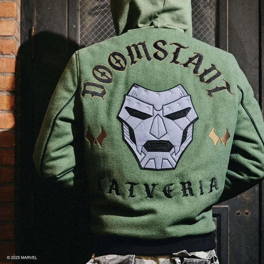 Person wearing a green varsity jacket with a large Doom Slayer helmet graphic on the back and curved text above standing in front of a brick wall and metal door