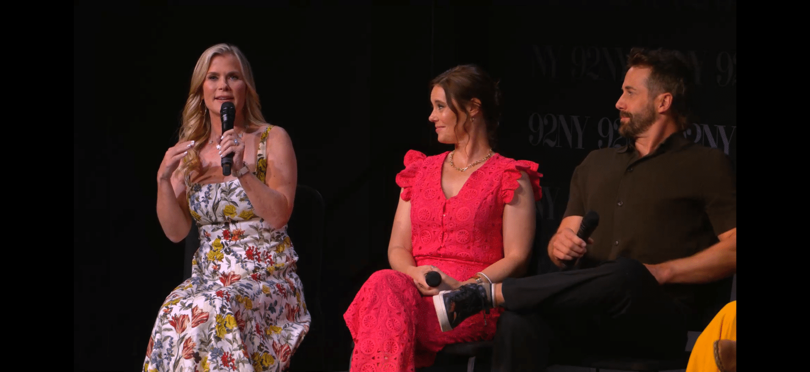 Three panelists on a stage a woman in a floral dress speaking into a handheld mic on the left a woman in a pink lace dress seated in the middle and a man in a dark shirt seated on the right