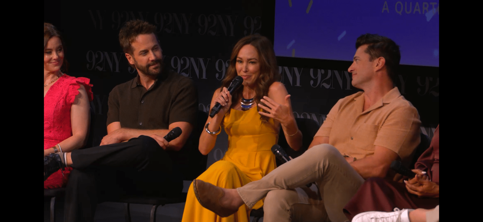 Woman in a mustard yellow dress speaks into a microphone on stage flanked by three seated panelists The background shows 92Y signage
