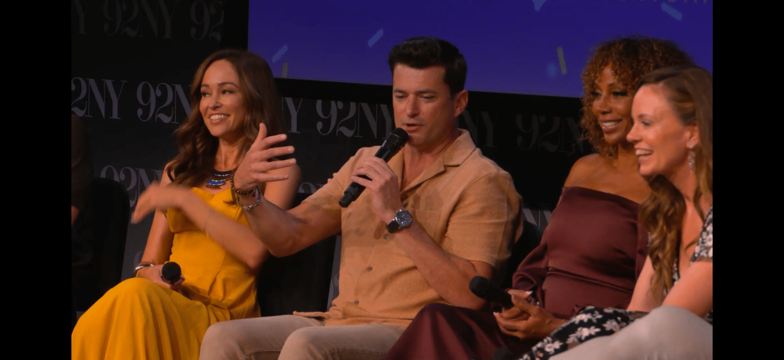 Panel of four adults on stage man in tan shirt speaks into a microphone while three women smile nearby seated in a row during a discussion