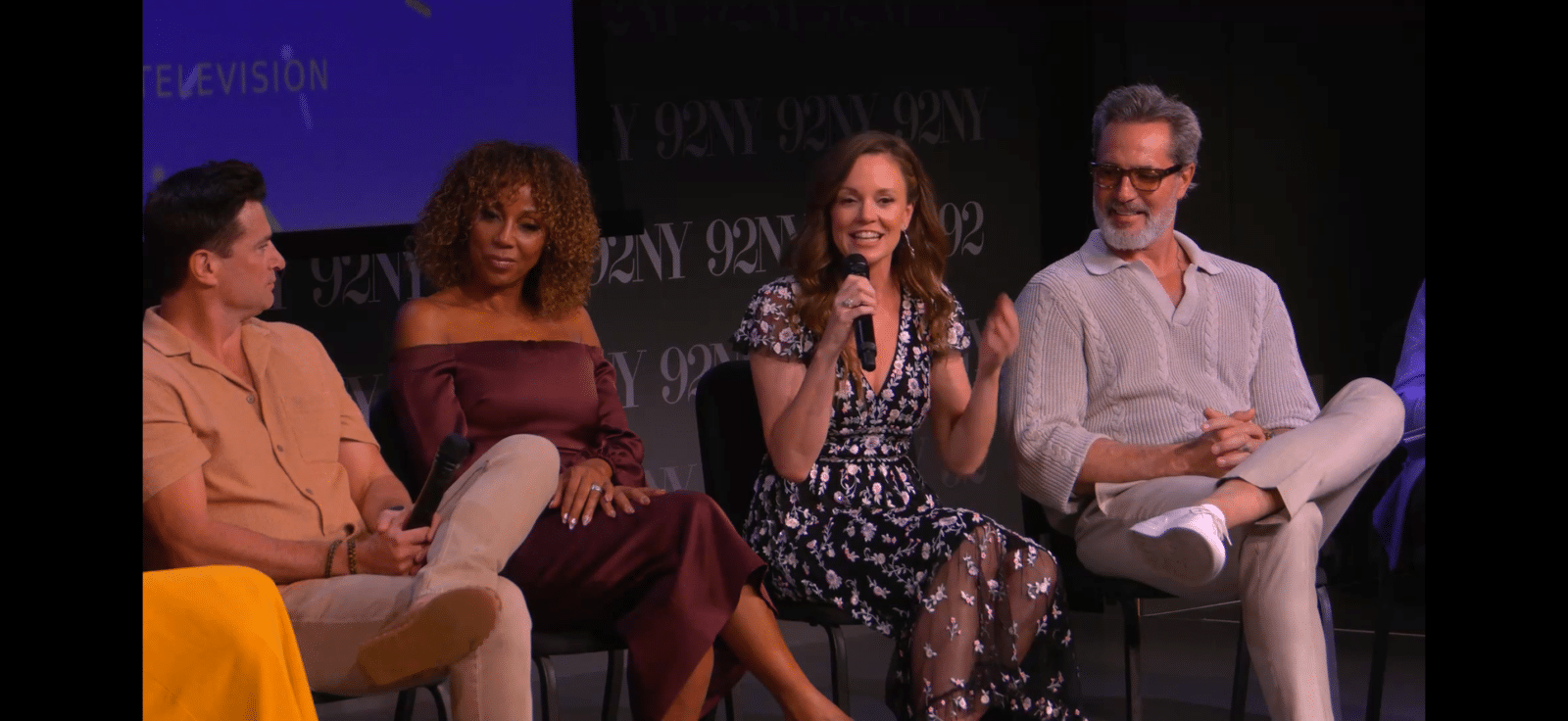 Panel of four people seated on a stage woman in floral dress speaking into a microphone Runner up on left and man with glasses on right listen