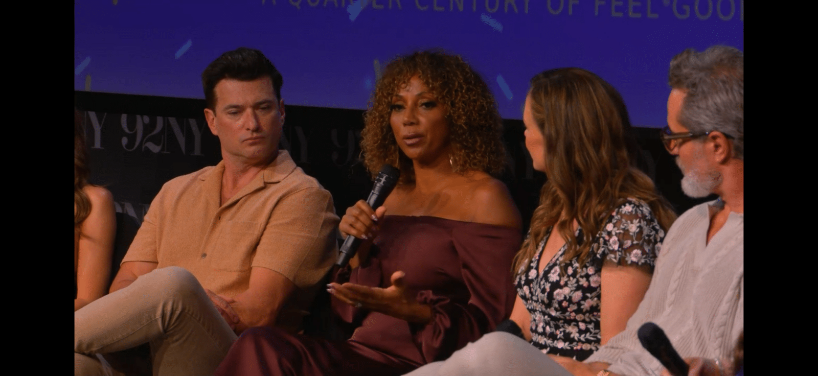 Panel of four people on stage woman center speaks into a microphone while others listen attentively