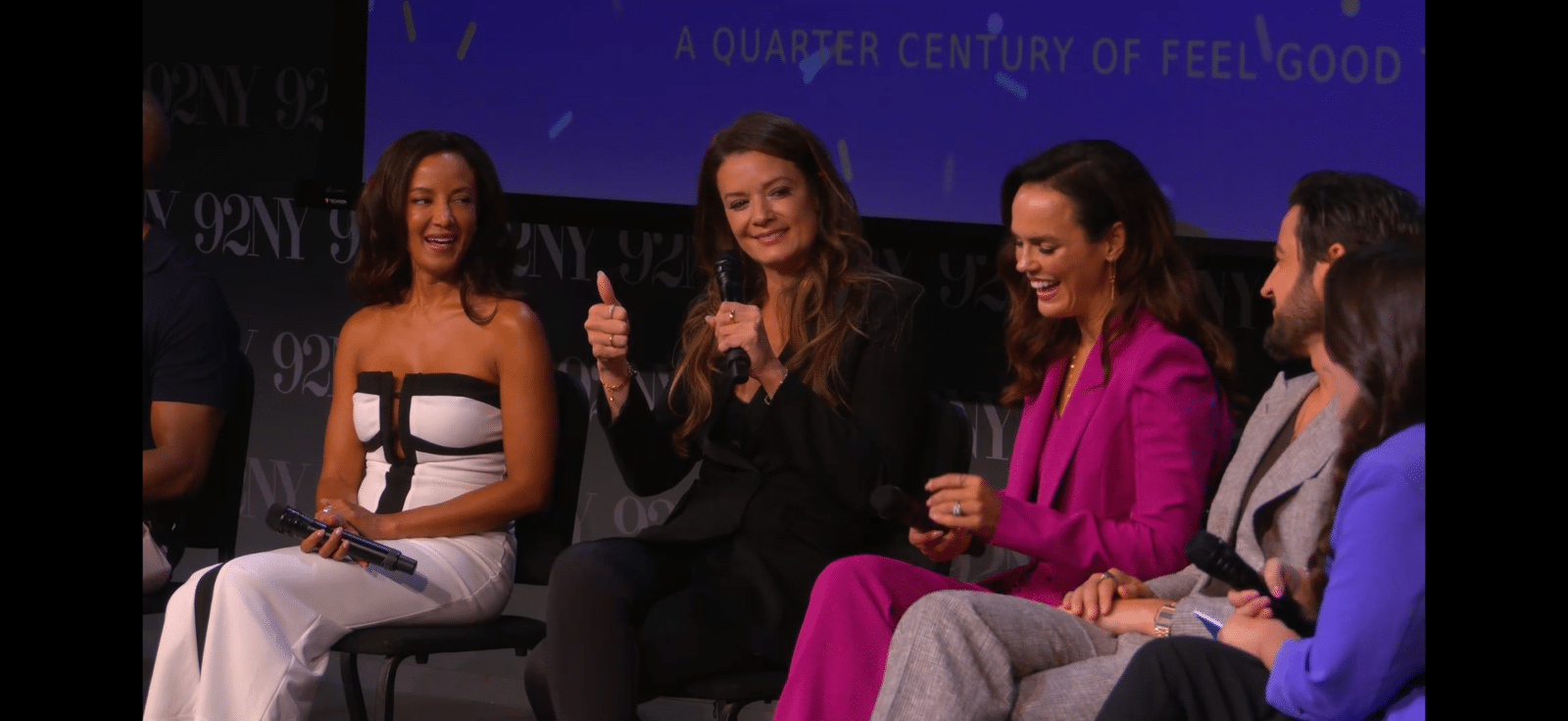 Panel of five women seated on stage during a discussion each holding a microphone smiling and chatting against a blue backdrop