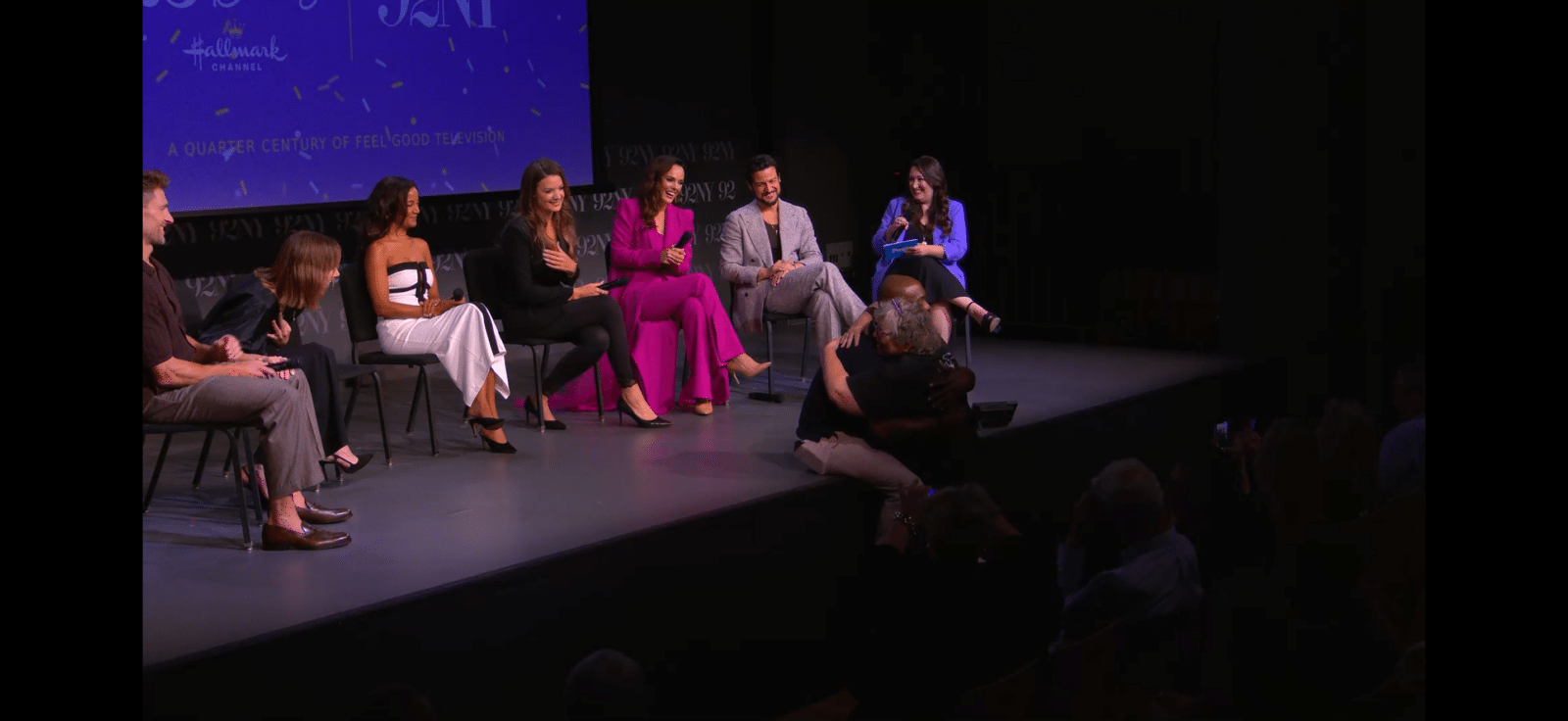Panel discussion on a stage seven people seated in a row with a blue backdrop and Hallmark Channel logo visible behind them
