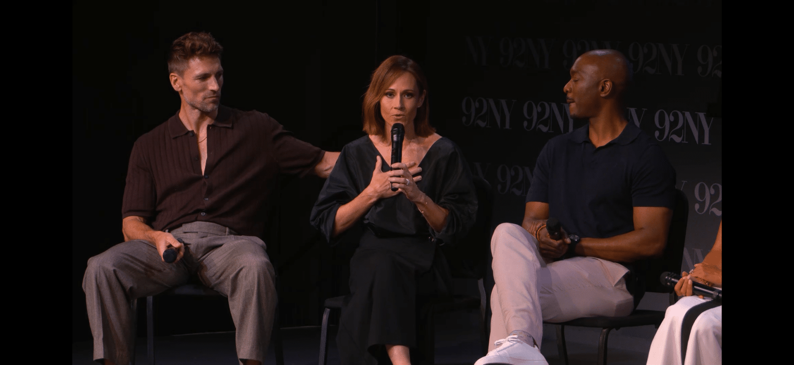 Three panelists seated on stage center woman speaks into a microphone while others listen each holding a mic against a dark backdrop