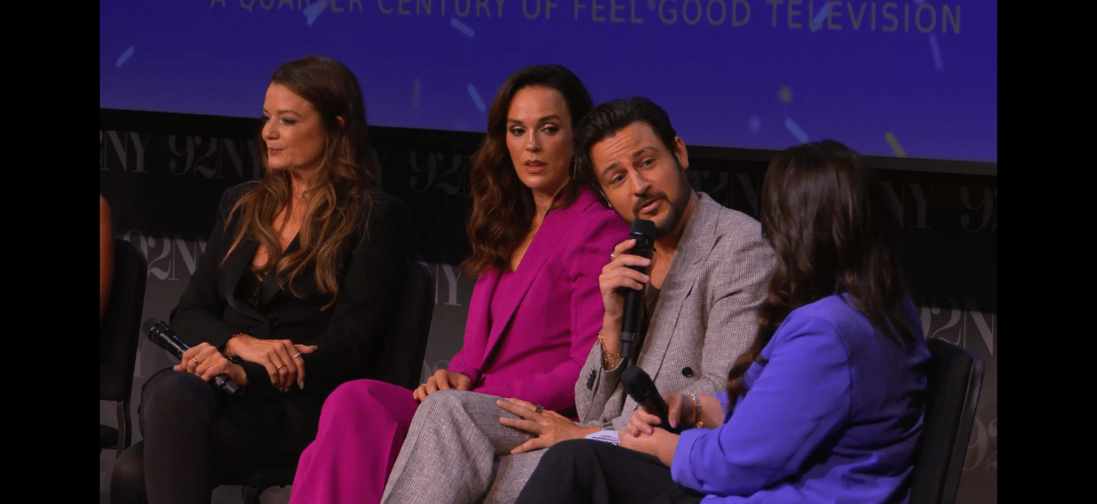 Panel of four people seated on stage during a discussion man in a gray suit speaks into a handheld microphone while others listen with a purple lit backdrop