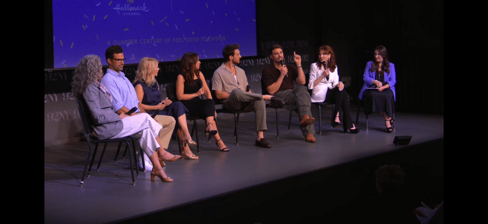 Panelists seated in a row on stage with microphones during a discussion blue backdrop behind them and a visible audience foreground