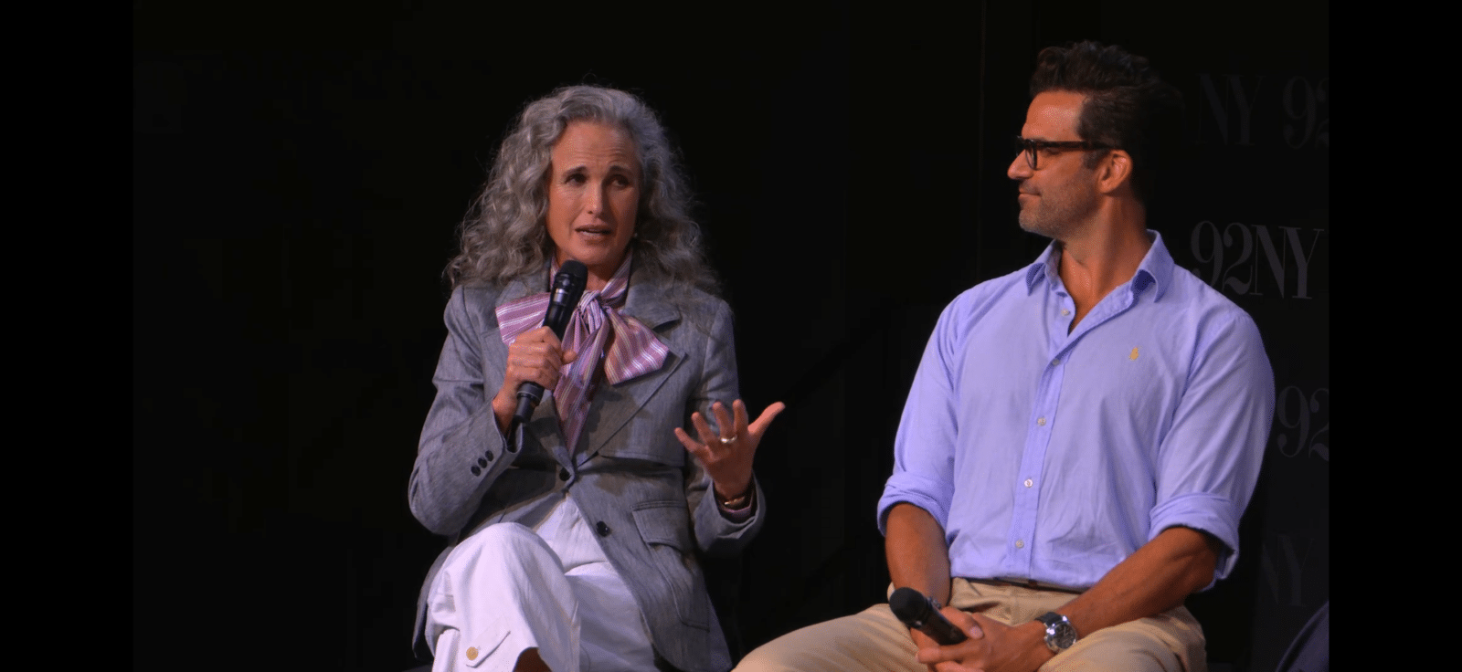 Two people sit on stage in conversation a woman with gray curly hair holding a microphone and speaking beside a man in a blue shirt listening attentively