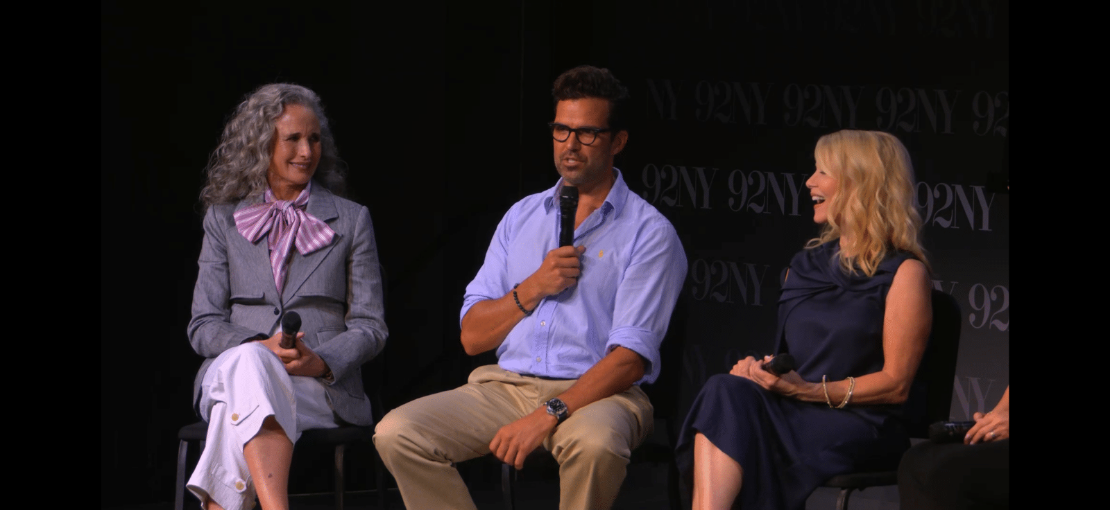 Three panelists on stage holding microphones left an older woman with gray hair in a gray suit and pink bow center a man in a light blue shirt right a blonde woman in a navy dress
