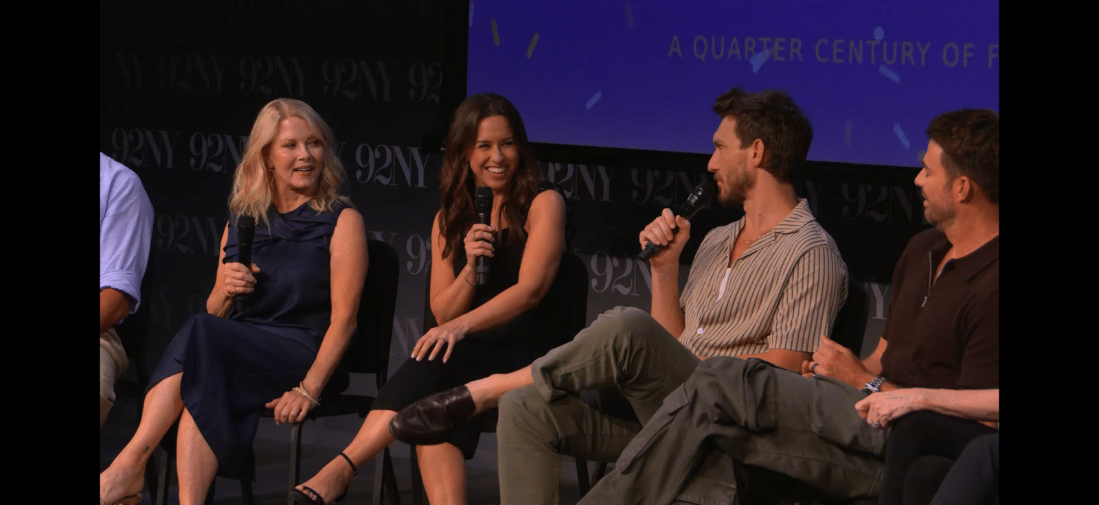 Five panelists sit on stage each holding a microphone mid conversation at a discussion event with a dark backdrop and a blue screen