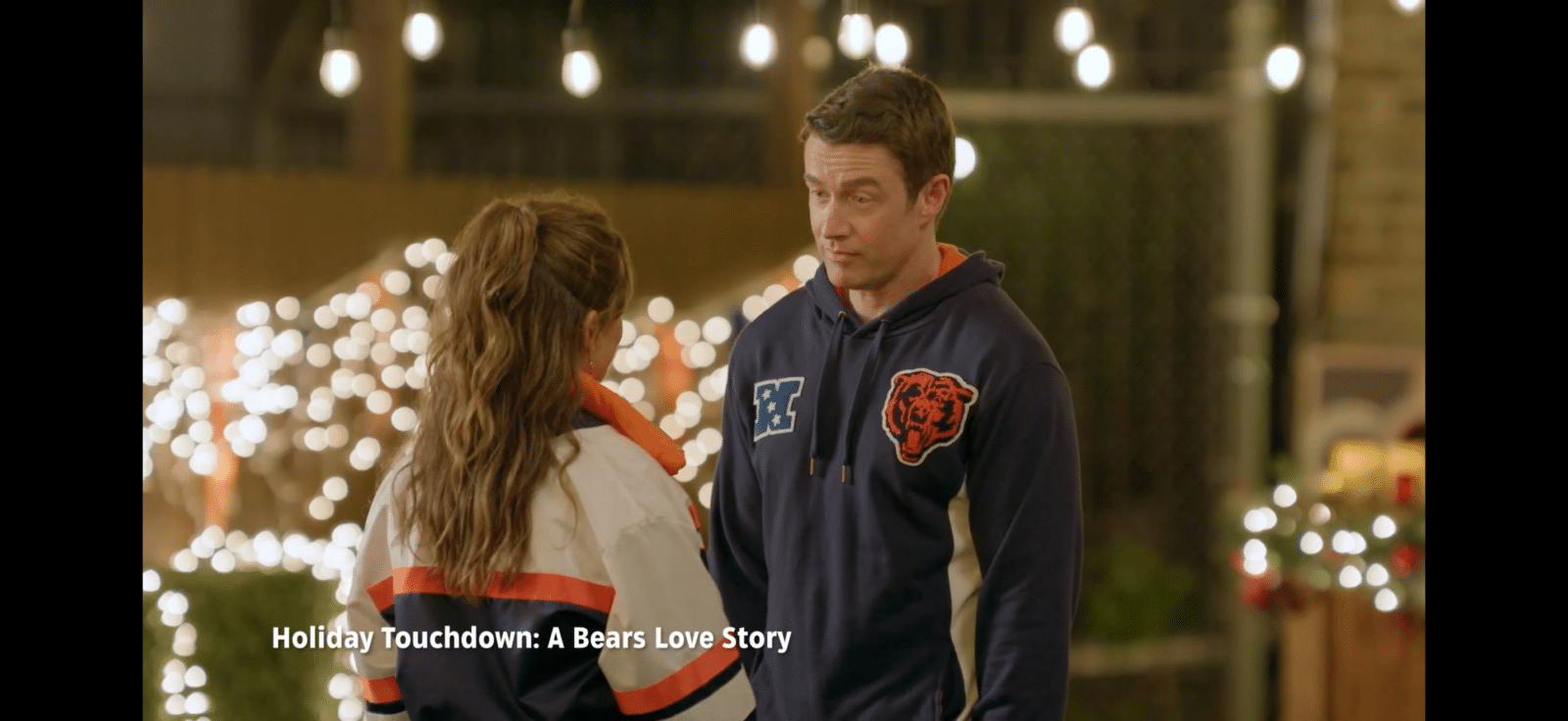 Two people face each other outdoors at night with festive string lights in the background man wears a Chicago Bears hoodie while talking to a woman in a white jacket with orange accents