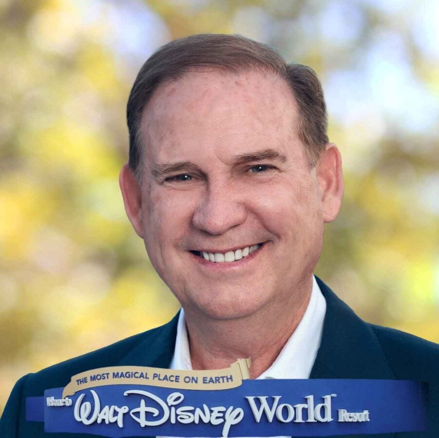 Smiling man in a dark suit holding a sign that says Walt Disney World Resort with a blurred yellow green background