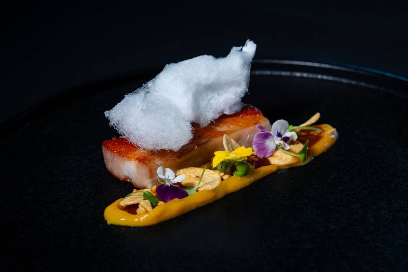 Seared fish fillet with a white foam garnish orange sauce and edible blossoms on a dark plate