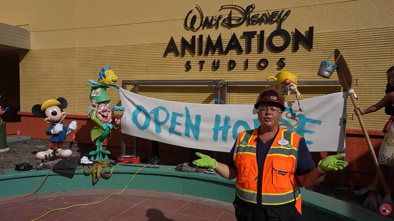 Outside Disney Animation Studios with a banner that reads OPEN HOUSE a worker in a bright orange safety vest and green gloves speaks to the camera as cartoon figures stand nearby
