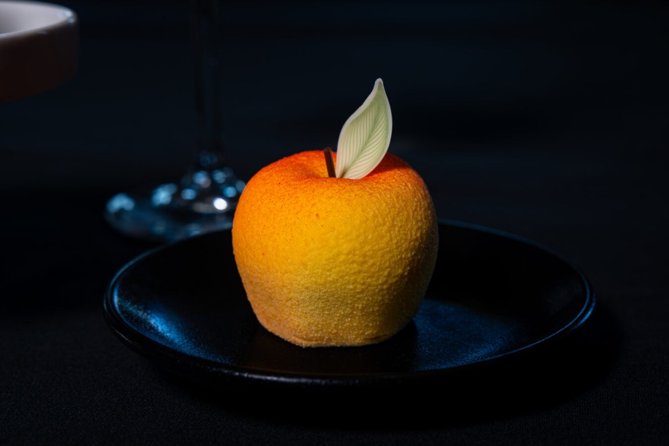 Orange dessert ball topped with a pale leaf garnish on a dark plate