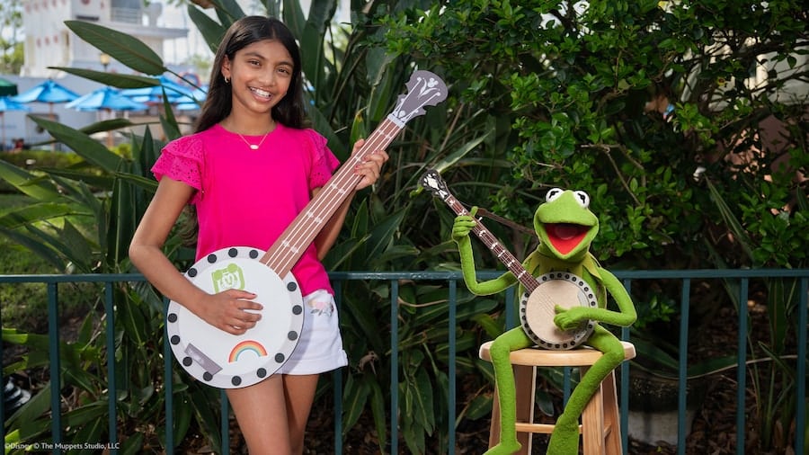 Girl in a pink shirt smiles while holding a decorated miniature ukulele a green frog puppet sits on a stool and plays a banjo beside her