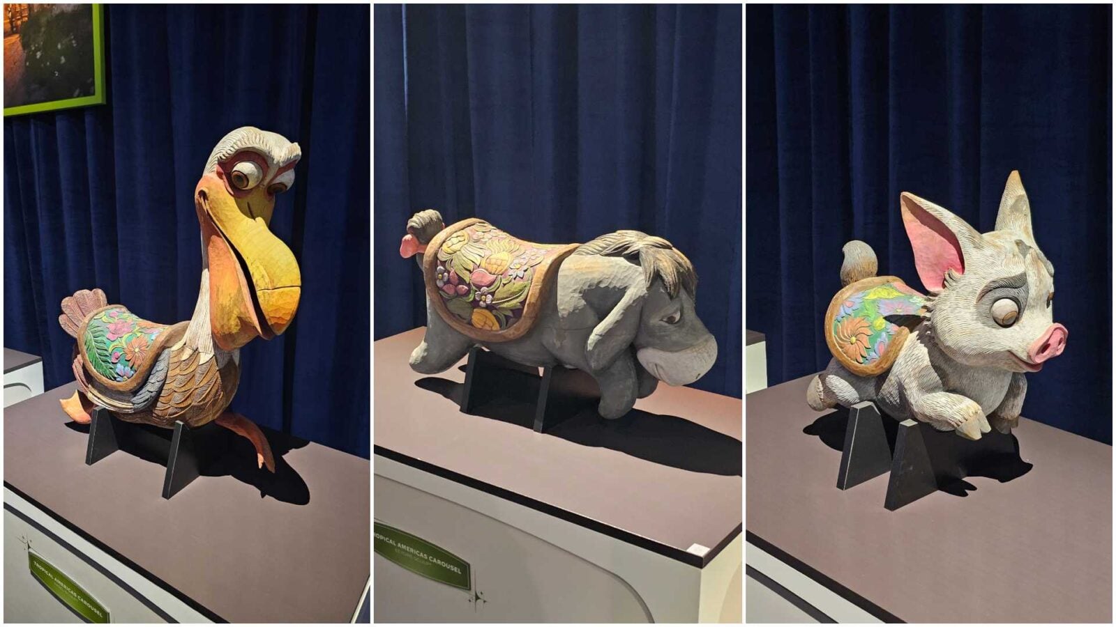 Triptych of carved wooden carousel animals on display a colorful duck on the left a decorated horse in the center and a pink eared pig on the right against a blue curtain