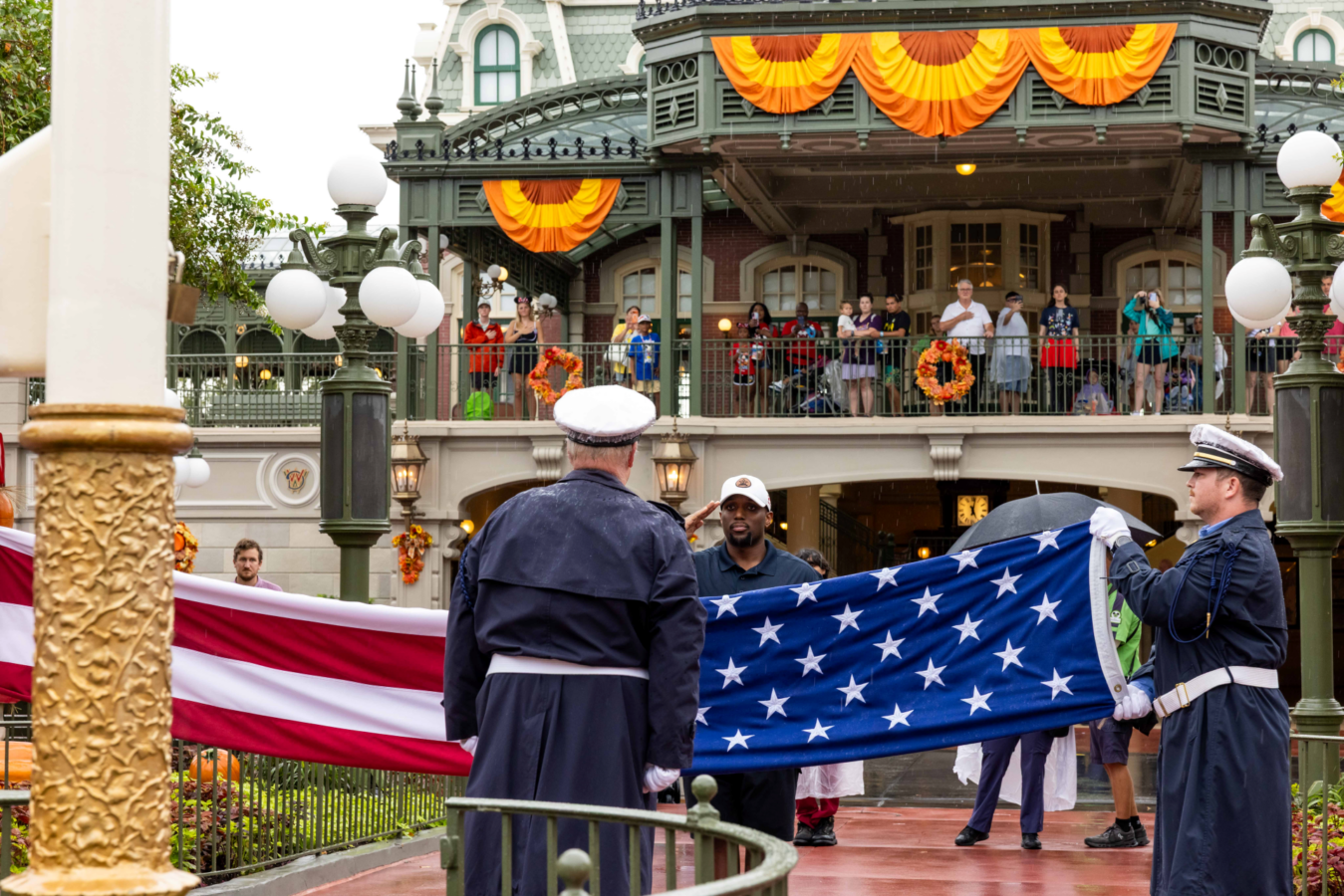 Guardians in navy uniforms hold a large American flag across a parade route as spectators watch from a decorated balcony above