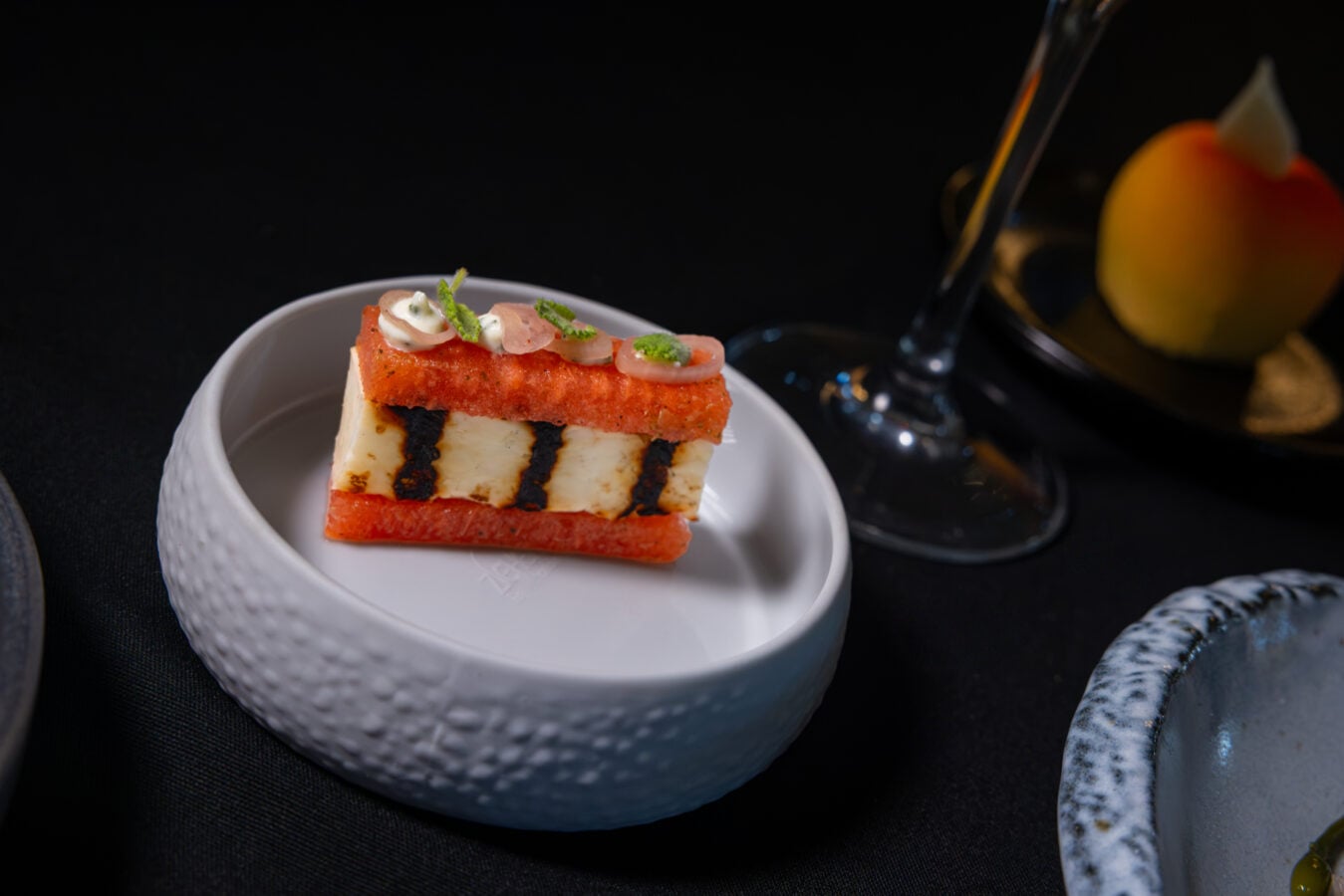 Slice of red cake with white cream layers on a white plate topped with small dollops of cream and green herbs against a dark background