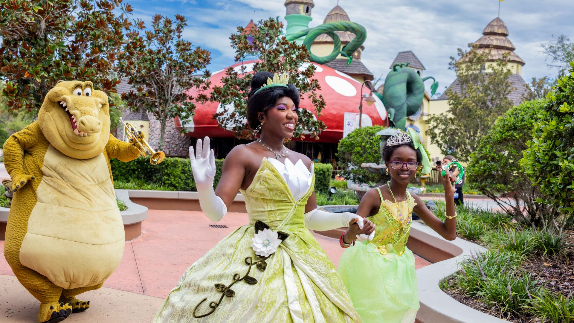 Yellow dinosaur mascot playing a saxophone beside two girls in green princess dresses at an outdoor theme park.