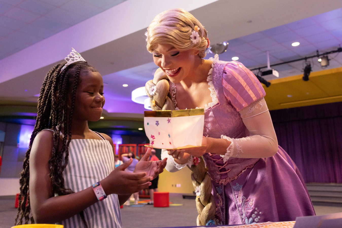 A smiling young girl in a striped dress receives a gift from a pink clad princess at a party or fair
