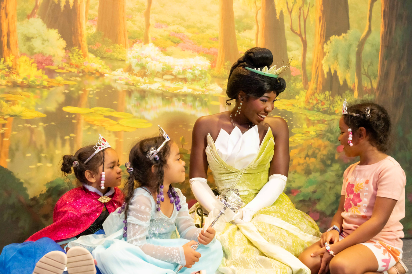 A woman in a yellow ball gown sits with young girls in princess costumes smiling in a colorful forest backdrop
