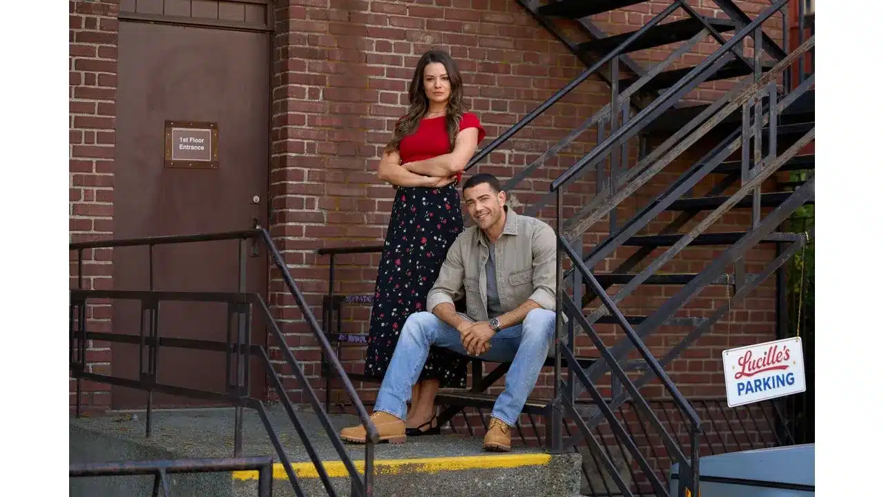 adty1 Fantasy Land News A woman in a red top and floral skirt stands with arms crossed behind a man seated on urban stairs outside a brick building