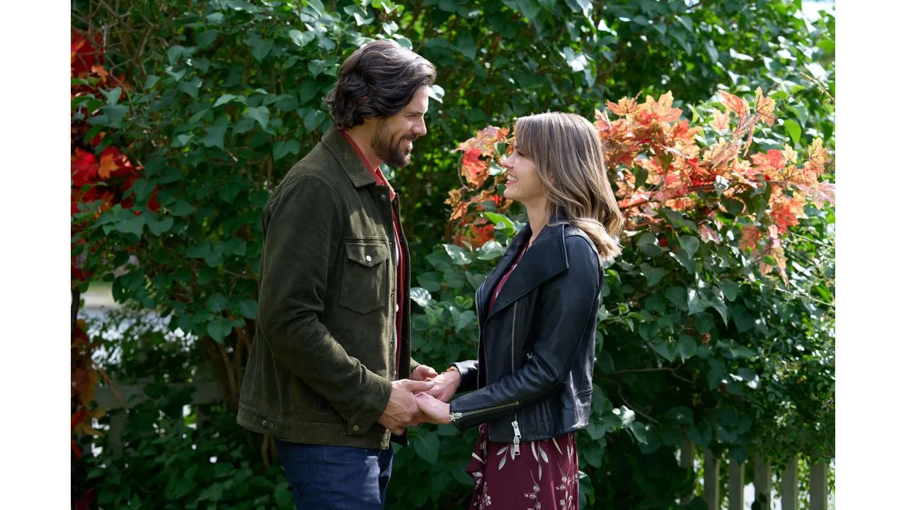 fall2 Fantasy Land News A couple stands outdoors holding hands and smiling at each other in front of green bushes with autumn red leaves behind them