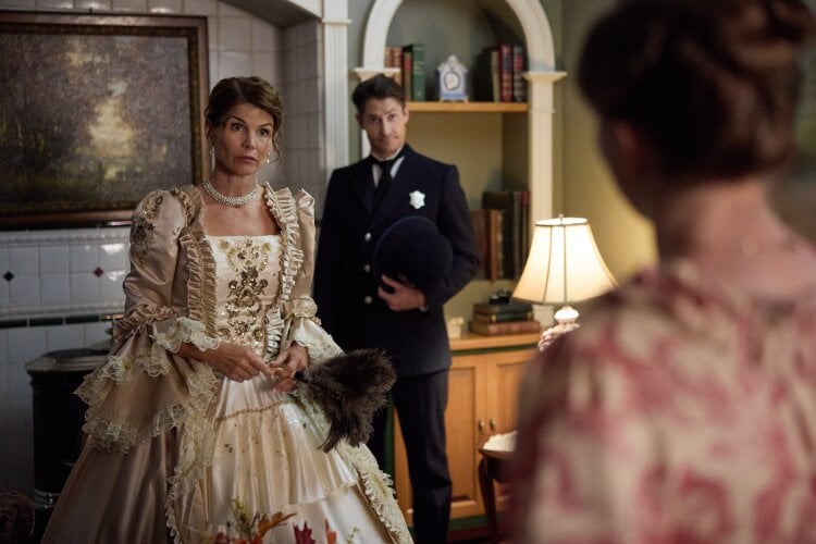 Elegant woman in a jeweled beige period gown holds a feathered fan as a man in a dark uniform stands in a vintage parlor behind her