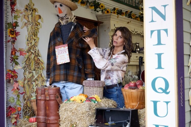Woman adjusts a life size scarecrow in a cottage style antique shop display with hay bales pumpkins and autumn decor visible