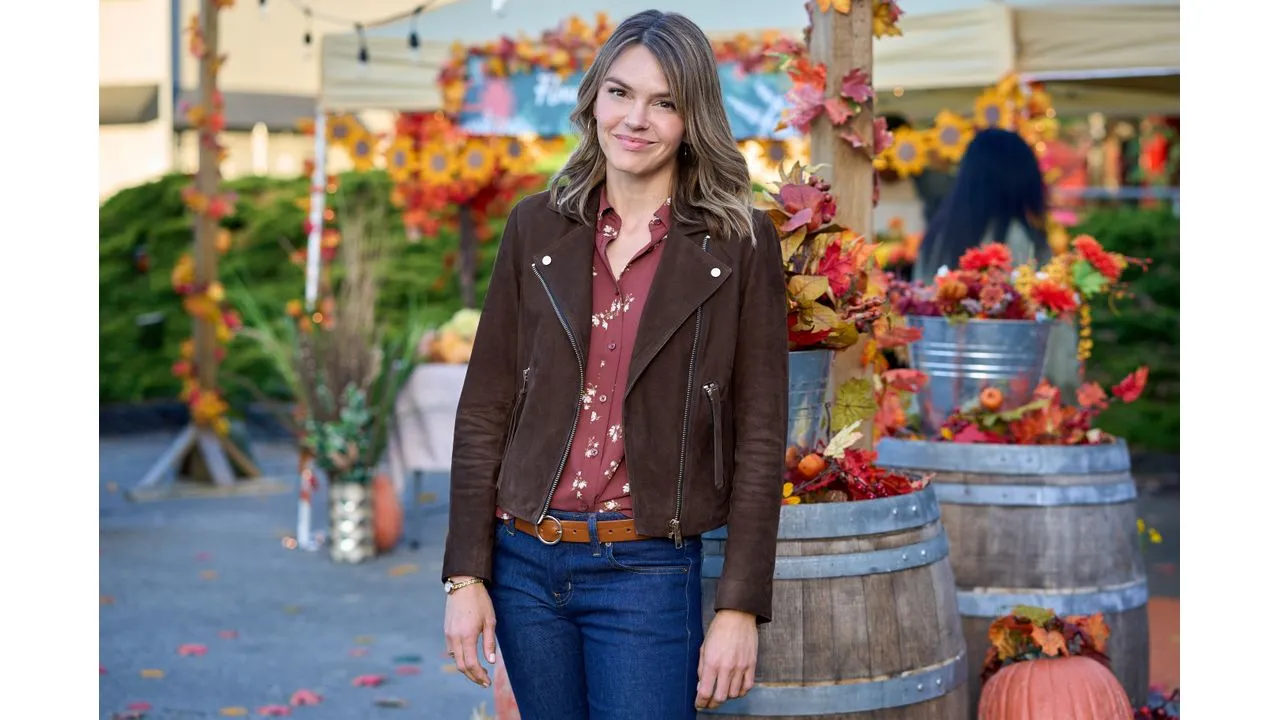 loved Fantasy Land News Woman in a brown suede jacket and red floral shirt posing at an autumn festival with pumpkins and barrel planters in the background