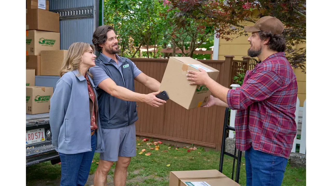 lovee Fantasy Land News Delivery driver hands a cardboard box to a man in a red plaid shirt in a suburban yard with a smiling couple and stacked boxes nearby