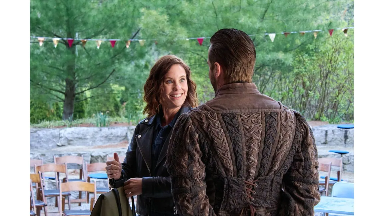 muchado3 Fantasy Land News Two adults converse outdoors at a casual event with colorful bunting and wooden chairs in a green park setting smiling at each other