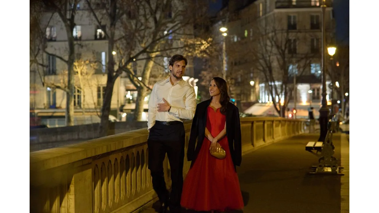paris4c Fantasy Land News Man in a white shirt and black pants walks beside a woman in a red dress with a black coat along a lit city promenade at night