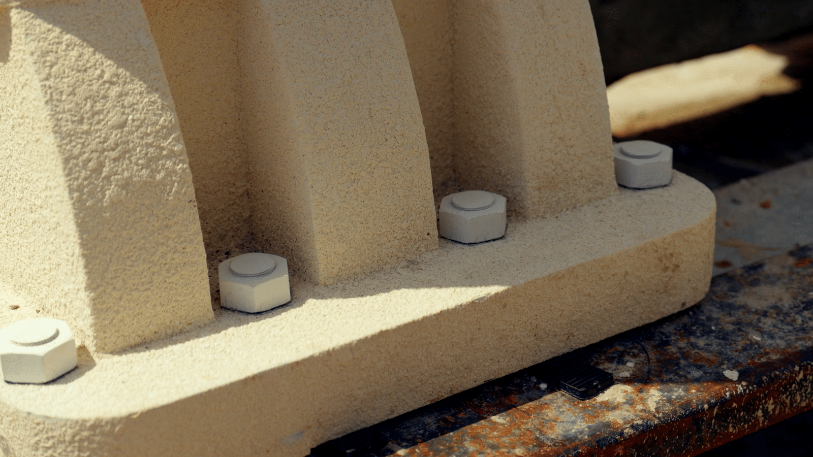 Close up of a beige concrete base with square bolt heads along its edge resting on a rusted metal surface