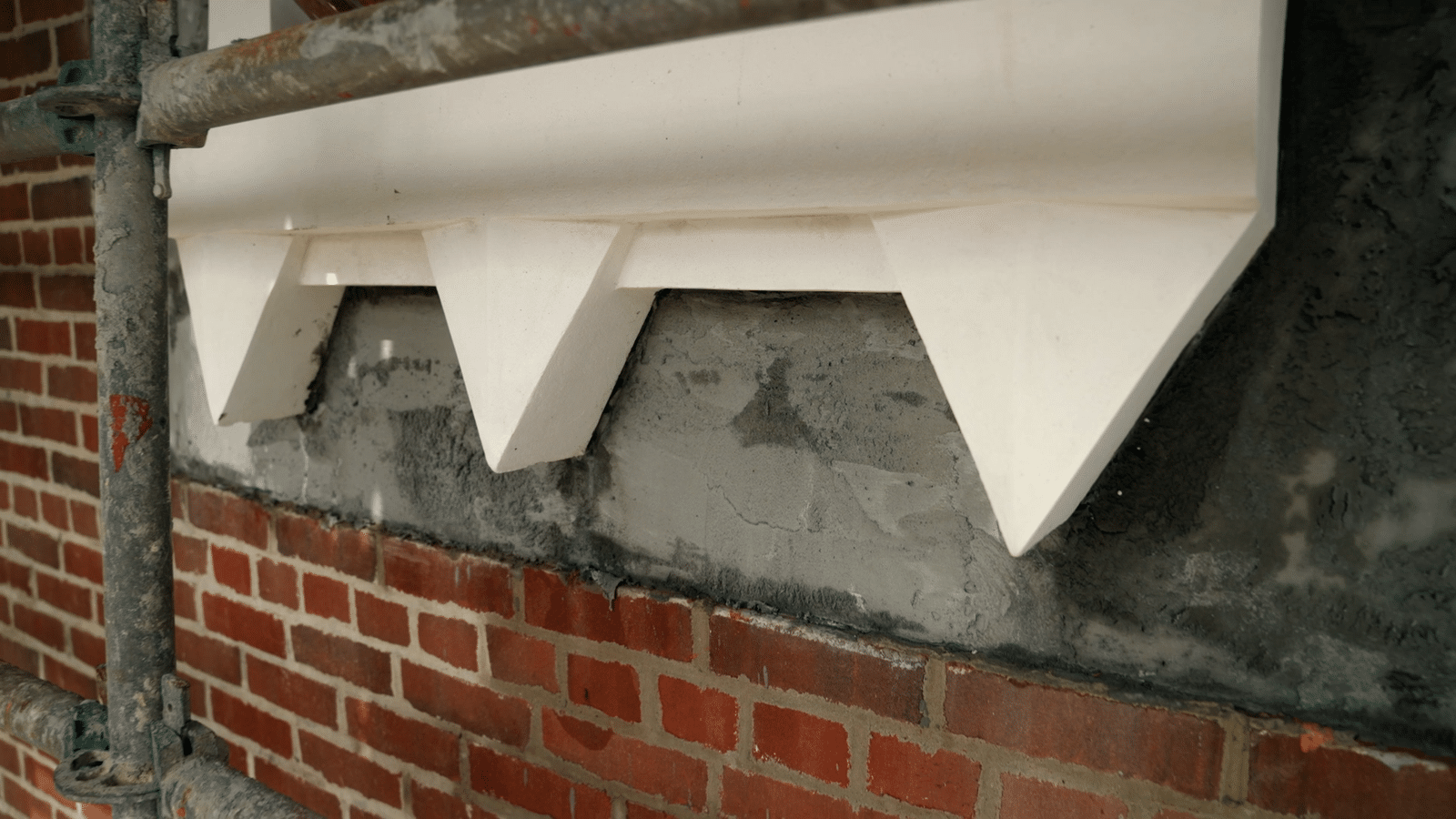 White decorative concrete brackets under a ledge on a red brick wall with a metal scaffold pipe in the foreground