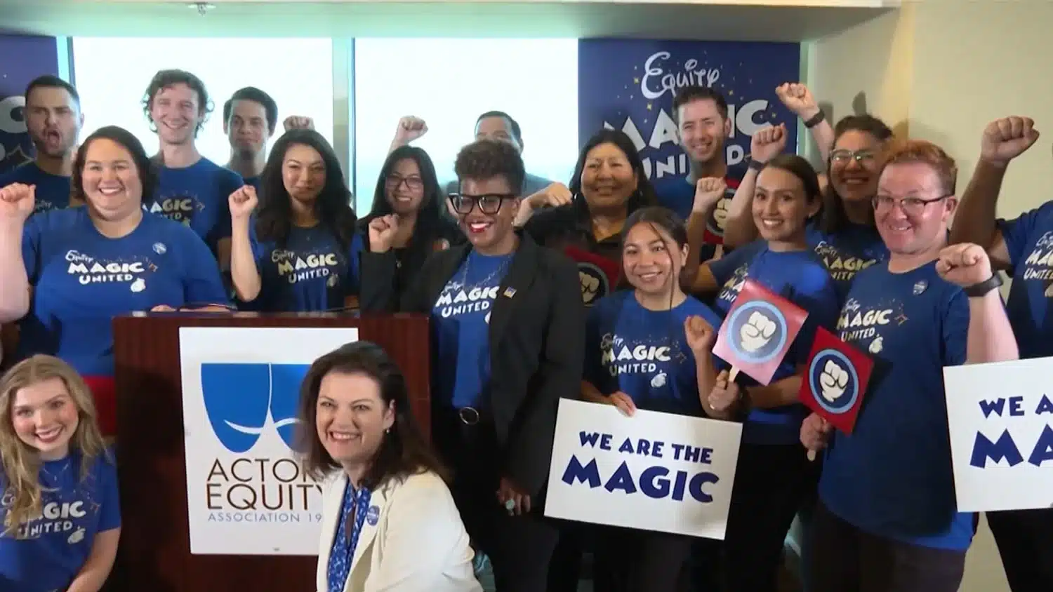 Group of diverse people in blue Magic United shirts cheer with raised fists holding signs like We Are the Magic at a team event Nearby podium bears a blue logo