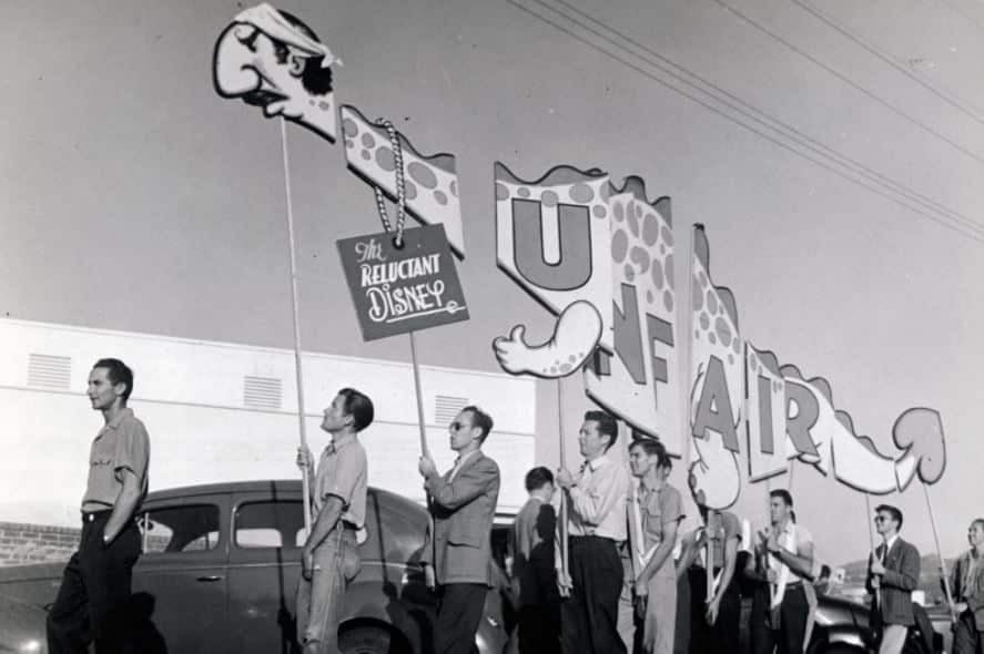 Black and white photo of a mid century parade men march beside a long float with oversized letters and a sign reading The Reluctant Disney A cartoon head decorates the float Car in the background