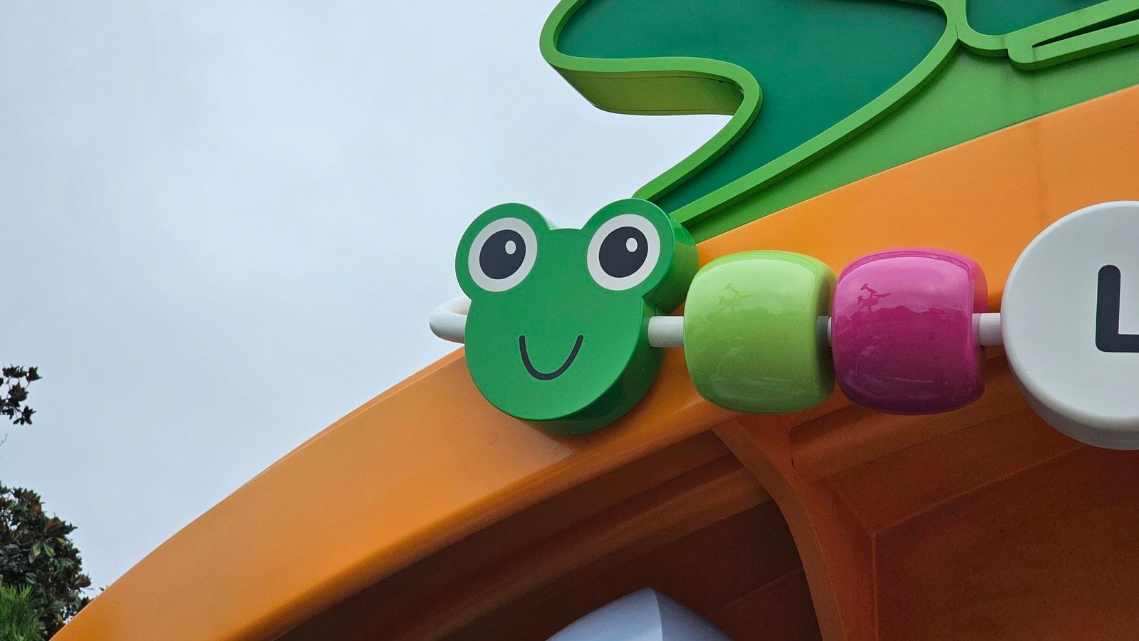 Close up of a green smiling cartoon frog head on a bright orange playground structure with oversized colorful beads and green trim