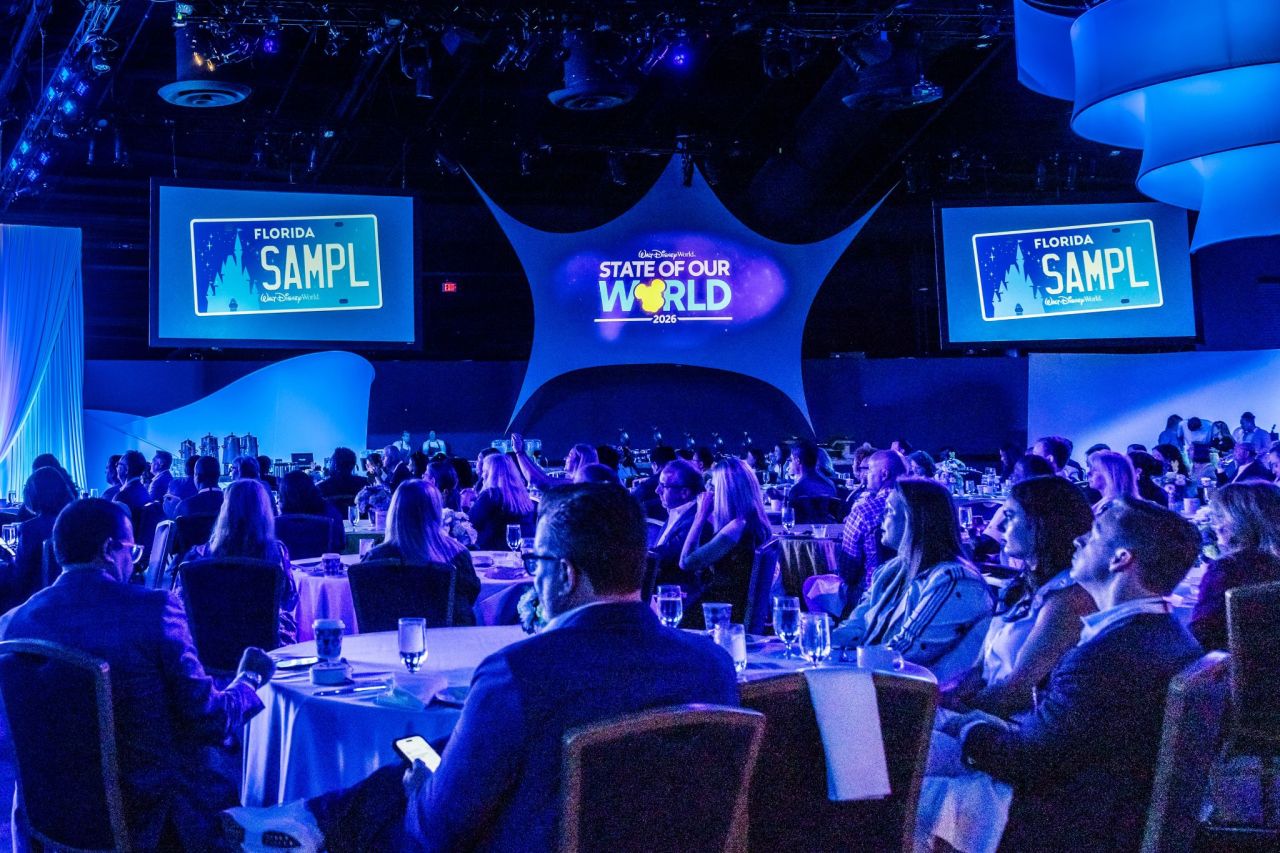 Audience seated at round tables in a blue lit conference hall watching a stage with a central screen reading State of Our World 2026 and side screens displaying graphics on either side of the stage conveying a formal event atmosphere across the room