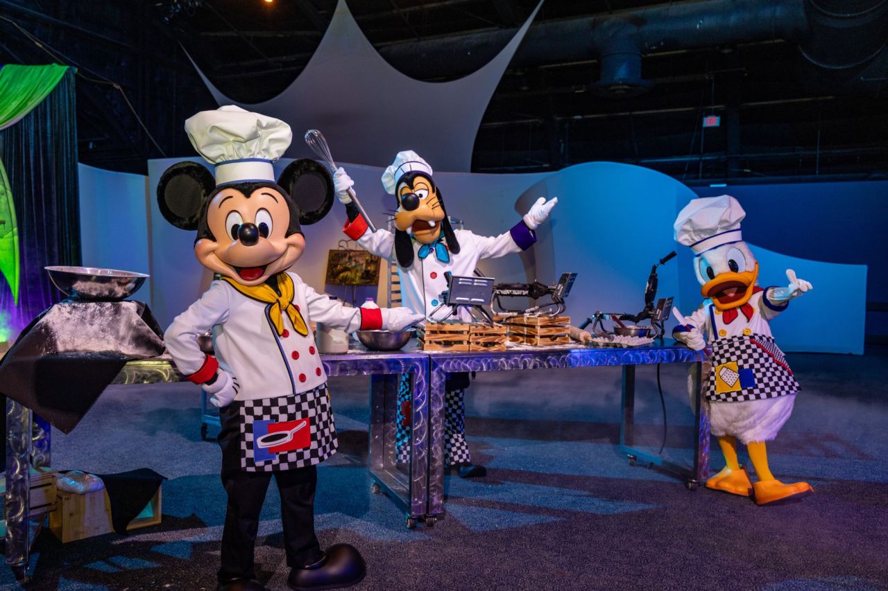 Mickey Mouse Goofy and Donald Duck in chef outfits posing at a kitchen prep table with cooking tools during a show