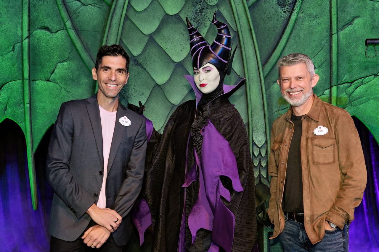 Three people pose with a Maleficent character in the center against a green dragon scale backdrop