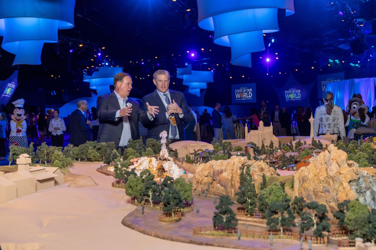 Two men in suits discuss a detailed miniature landscape model at a blue lit event hall