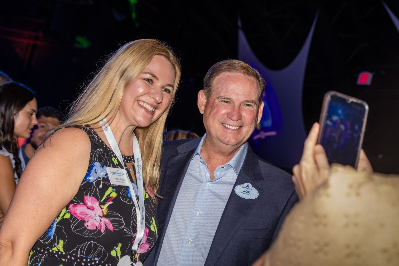 Two smiling attendees posing for a selfie at an event woman in a floral dress with a lanyard and man in a blazer with a name badge on his lapel and a blue shirt